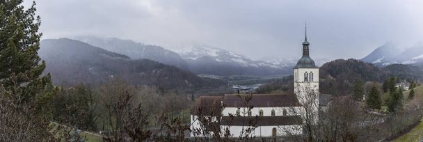 táj, ahol a templom látható a közepén a hegy, egy erdőben sötét színek, felhős téli nap köd, havas hegyek - Fotó, kép