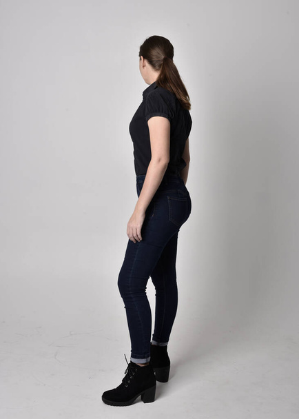 Porträt eines hübschen, brünetten Mädchens mit Pferdeschwanz, das Jeansstiefel und eine schwarze Bluse trägt. Stehende Pose in voller Länge im Seitenprofil, auf Studiohintergrund. - Foto, Bild