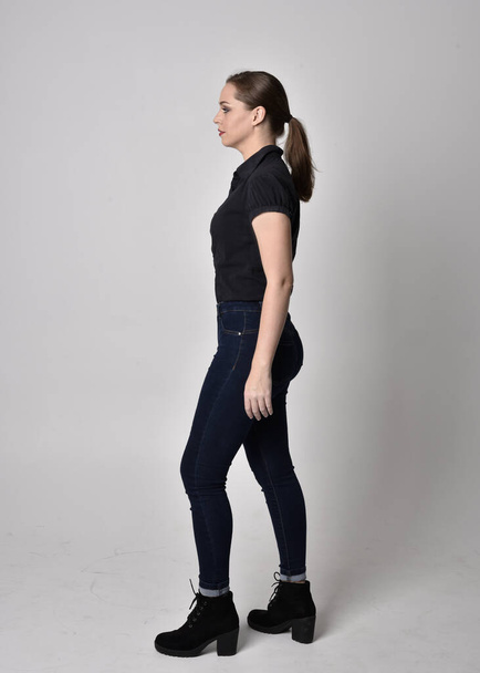 Portrait of a pretty brunette girl with ponytail, wearing jeans boots and a black blouse.  full length standing pose in side profile,  on a studio background. - Photo, Image