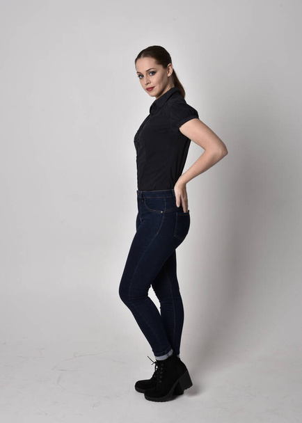 Portrait of a pretty brunette girl with ponytail, wearing jeans boots and a black blouse.  full length standing pose in side profile,  on a studio background. - Fotografie, Obrázek