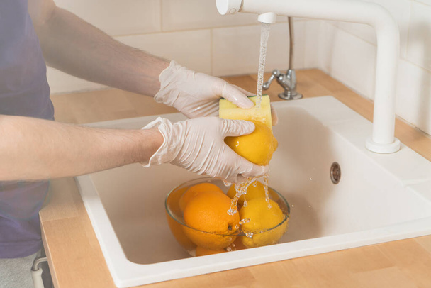 Mężczyzna myje cytrynę detergentem w rękawiczkach. Ręce w rękawiczkach jednorazowego użytku zbliżenie umyć cytrynę pod bieżącą wodą. - Zdjęcie, obraz