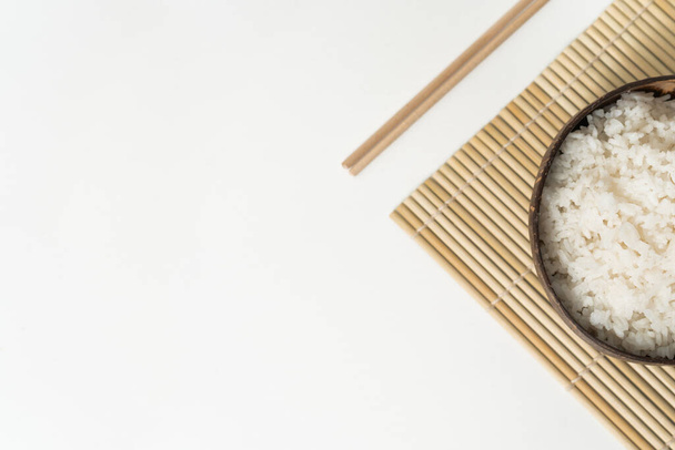Arroz branco em uma tigela de coco em um fundo branco. Foto minimalista com pauzinhos de arroz e bambu
 - Foto, Imagem