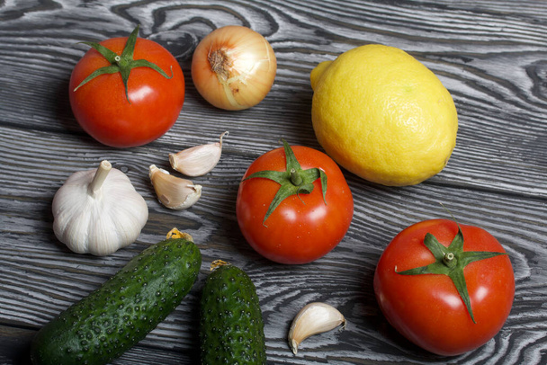 Tomatoes with green ponytails, onion, lemon and garlic head. Two green prickly cucumbers with yellow flowers. They lie on the surface of brushed pine boards. - Photo, Image