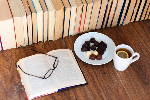 Open textbook, glasses, cup of tea and chocolate candies on white plate, stack of old book on wooden table, education concept background, many books piles with copy space for text - Photo, Image