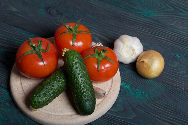 Cucumbers, tomatoes, onions and garlic on a cutting board. Tomatoes with green ponytails. Cutting board lies on the surface of brushed pine boards. - Photo, Image