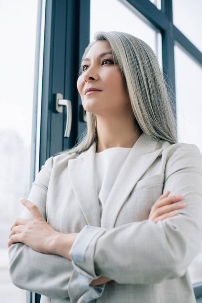 confident asian businesswoman with grey hair and crossed arms in office - Фото, изображение