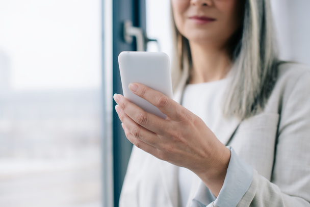 vista recortada de la mujer de negocios con el pelo gris usando smartphone en la oficina
  - Foto, Imagen