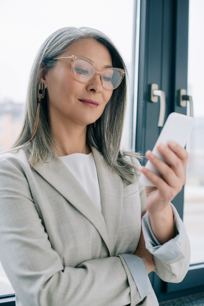 attractive asian businesswoman in eyeglasses with grey hair using smartphone in office  - Фото, изображение