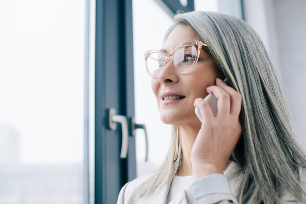 professionista asiatica donna d'affari in occhiali con i capelli grigi parlando su smartphone in ufficio
  - Foto, immagini