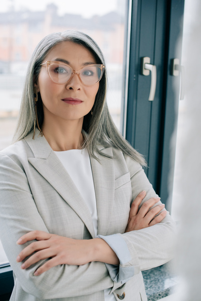 confident asian businesswoman with crossed arms in grey suit and eyeglasses in office - Фото, изображение