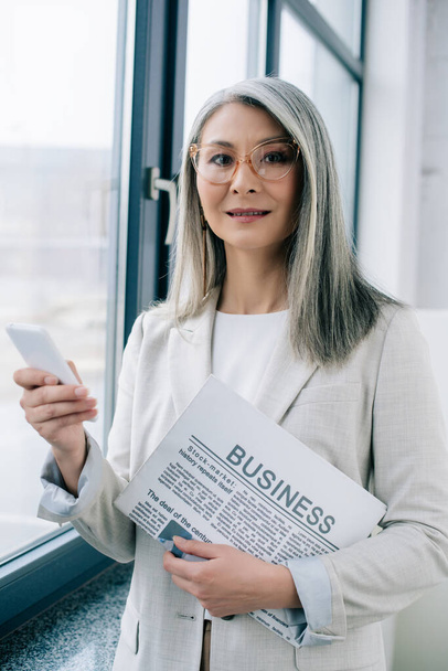 beautiful asian businesswoman in eyeglasses using smartphone and holding business newspaper in office - Фото, изображение