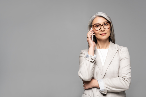 asian businesswoman in eyeglasses with grey hair talking on smartphone isolated on grey - Фото, зображення