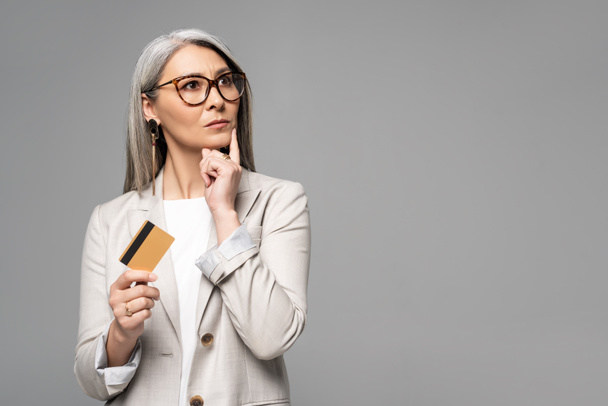 mooie doordachte aziatische zakenvrouw met grijs haar houden credit card geïsoleerd op grijs - Foto, afbeelding
