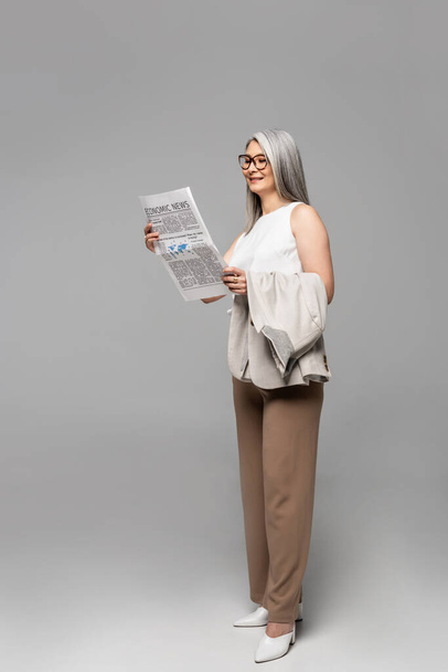 attractive asian businesswoman in eyeglasses reading business newspaper on grey - Фото, зображення