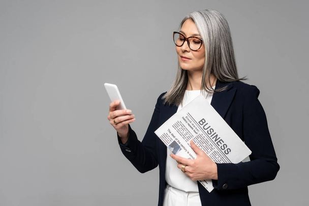 confident asian businesswoman in eyeglasses using smartphone and holding business newspaper isolated on grey - Фото, изображение