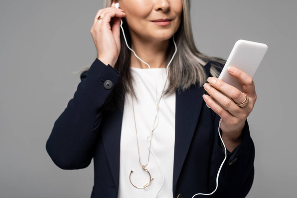 cropped view of businesswoman with grey hair listening music with earphones and smartphone isolated on grey - Fotó, kép