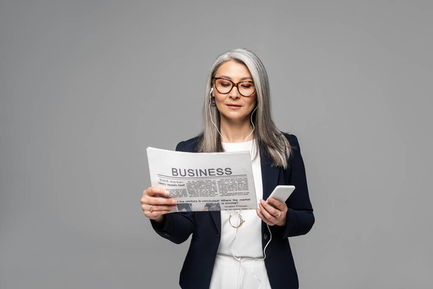 attractive asian businesswoman in eyeglasses listening music with earphones and smartphone while reading business newspaper isolated on grey - Foto, afbeelding