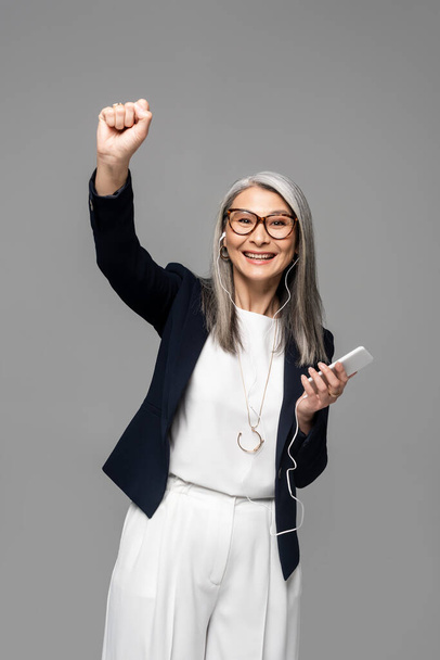 excited asian businesswoman with grey hair listening music with earphones and smartphone isolated on grey - Valokuva, kuva