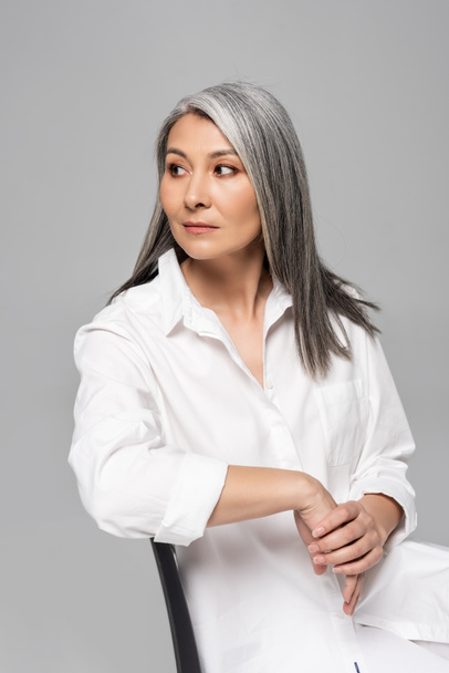 confident asian woman with grey hair sitting on chair isolated on grey - Zdjęcie, obraz