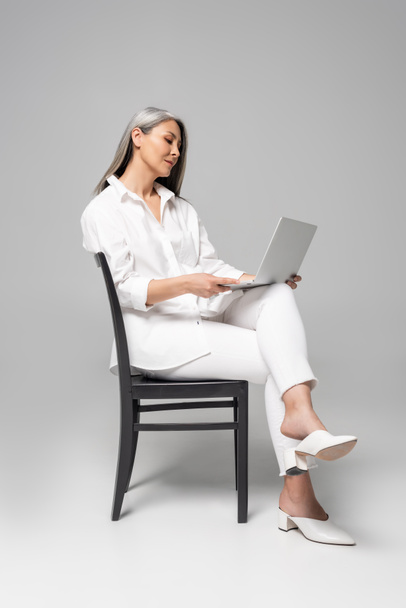 adulto atraente asiático mulher com cabelos grisalhos sentado na cadeira e usando laptop no cinza
 - Foto, Imagem