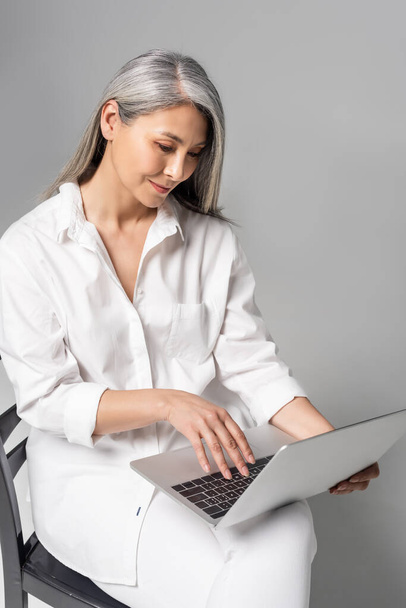smiling asian woman with grey hair sitting on chair and using laptop on grey  - Zdjęcie, obraz
