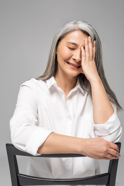 beautiful laughing asian woman with grey hair sitting on chair isolated on grey - 写真・画像