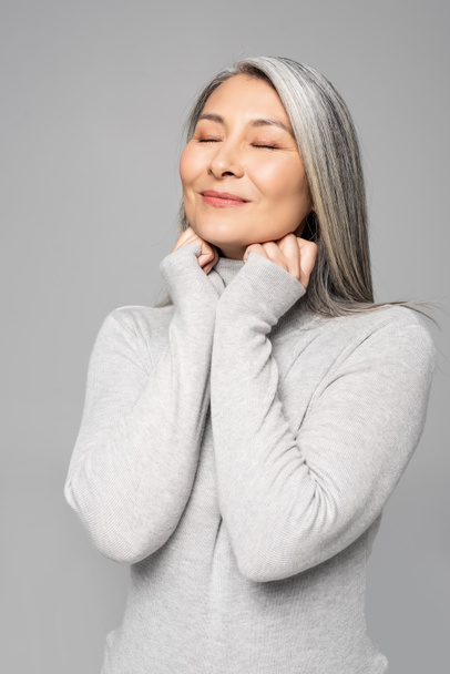 lächelnde asiatische Frau im Rollkragen mit grauen Haaren und geschlossenen Augen isoliert auf grau - Foto, Bild
