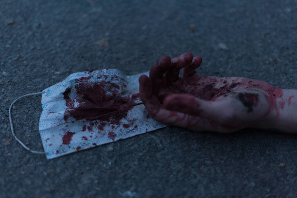 A medical bloodied mask lies on the ground with a bloody hand reaching out. The concept of coronavirus and the apocalypse resulting from this disease has spread throughout the world. Horror. - Fotografie, Obrázek