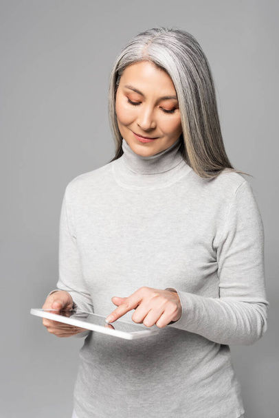 happy adult asian woman in turtleneck using digital tablet isolated on grey  - Foto, imagen