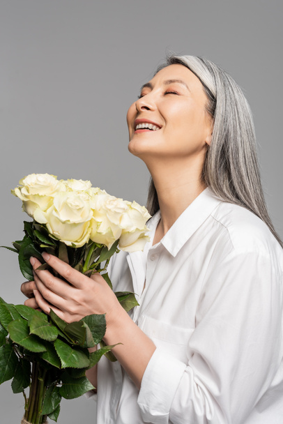 adult cheerful asian woman with grey hair holding bouquet of white roses isolated on grey - Foto, Bild