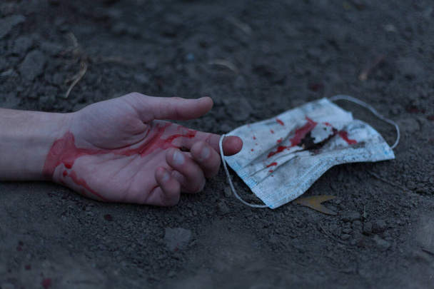 A medical bloodied mask lies on the ground with a bloody hand reaching out. The concept of coronavirus and the apocalypse resulting from this disease has spread throughout the world. Horror. - Фото, изображение