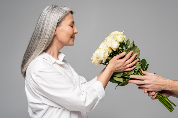 positive adult asian woman with grey hair in white shirt taking bouquet of white roses from man isolated on grey - Fotó, kép