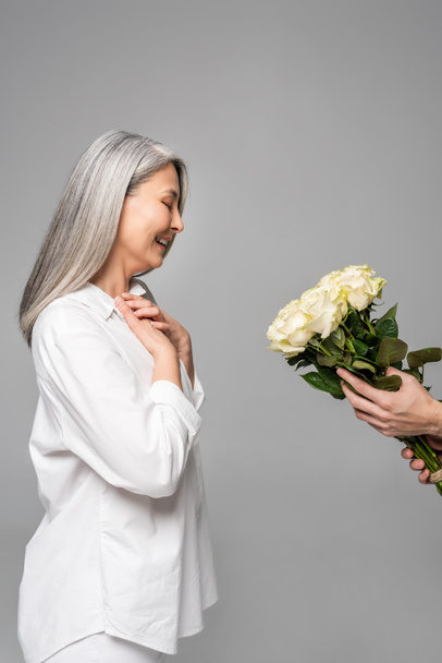 cheerful adult asian woman with grey hair in white shirt taking bouquet of white roses from man isolated on grey - Zdjęcie, obraz