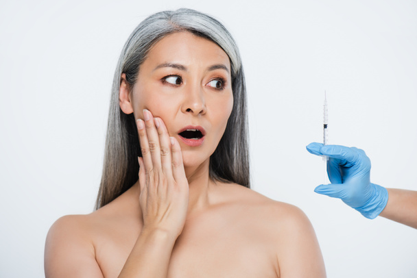 schockierte nackte asiatische Frau und Arzt in Latexhandschuhen mit Spritze und Schönheitsinjektion isoliert auf grau - Foto, Bild