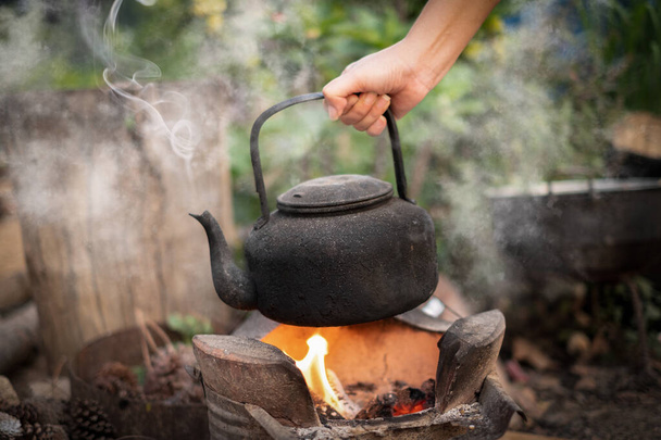 Zamknij dłoń trzymając wrzącą wodę stary czajnik na ogniu z piecem węglem drzewnym na rozmytym tle - Zdjęcie, obraz