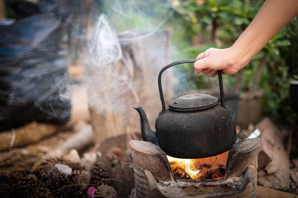 Cerca de la mano sosteniendo hervir agua vieja hervidor de agua en el fuego con una estufa de carbón al fondo borroso
 - Foto, Imagen