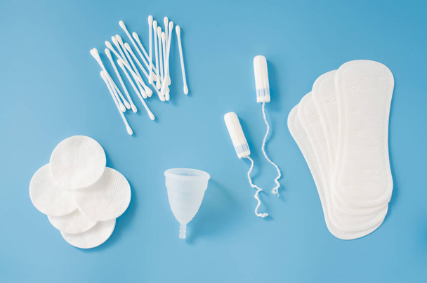 Feminine hygiene accessories. Concept of feminine hygiene during menstruation. Sanitary pad, menstrual cup and tampons, cotton buds and cotton pads on blue background. Flat lay, top view - 写真・画像
