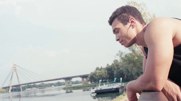 Sportowiec ze słuchawkami opartymi na ogrodzeniu, uśmiechnięty i patrzący na rzekę - Materiał filmowy, wideo