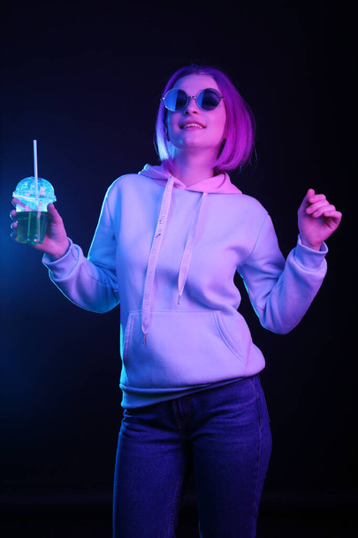 Woman in neon light and sunglasses. Dancing in a club and holding a glass in his hand with a drink, a cocktail. Night life. Generation Y, millennials. - Fotografie, Obrázek