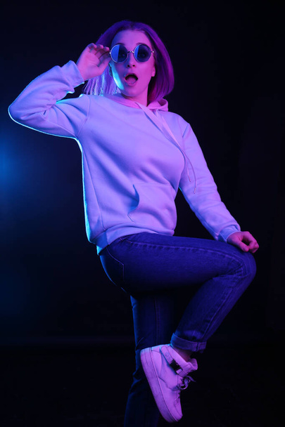 A woman with colored hair, an informal female drowns on a black background in a club, in neon lights and sunglasses, dancing. The girl is dressed in a lilac sweatshirt, sweater. Night life. - Photo, Image
