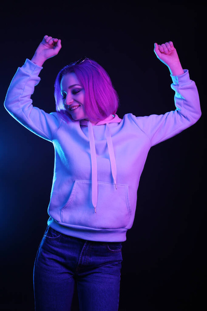 A woman with colored hair, an informal woman drowns on a black background in a club, in neon light. The girl is dressed in a lilac sweatshirt, sweater. Night life. - Foto, Imagen
