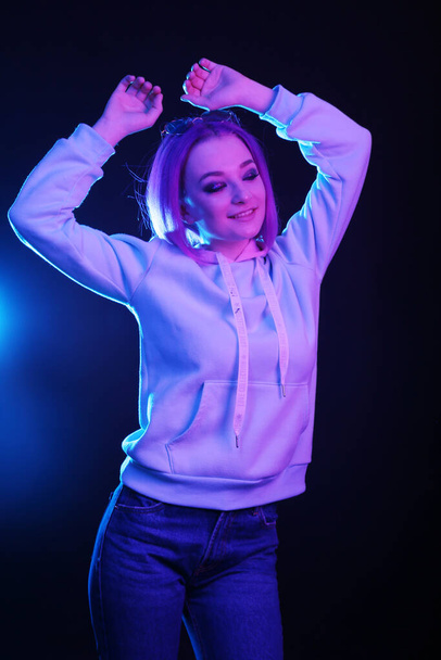 A woman with colored hair, an informal woman drowns on a black background in a club, in neon light. The girl is dressed in a lilac sweatshirt, sweater. Night life. - Zdjęcie, obraz