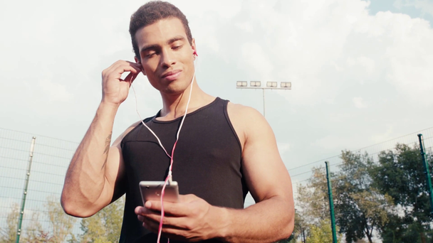 Vista de bajo ángulo del deportista poniendo auriculares en las orejas y sonriendo en la calle
 - Metraje, vídeo
