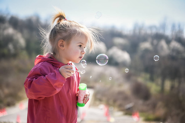 Двухлетняя кавказская девочка, пускающая мыльные пузыри на открытом воздухе в течение дня - счастливое беззаботное детство
 - Фото, изображение