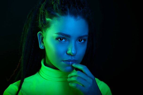 Sexy portrait of a young woman, in a neon blue and green light. Girl with dreadlocks in a nightclub, sexy turtleneck, radiant makeup with highlighter. Glitter skin. Clubber, night life concept - Fotó, kép