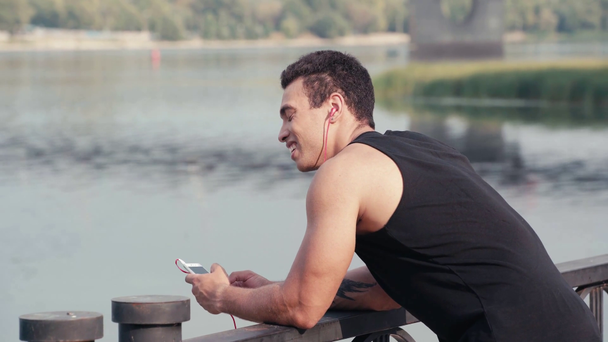 Sportler mit Kopfhörern plaudert auf Smartphone, lächelt und lehnt sich an Zaun - Filmmaterial, Video