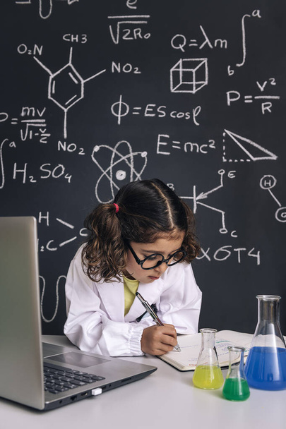 little scientist girl with glasses studying with a computer and taking notes, on school blackboard background with science formulas, back to school and successful female career concept, vertical photo - Photo, Image