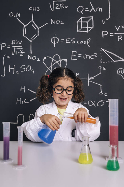 little scientist girl with glasses in lab coat mixing chemical liquids in flasks, on blackboard background with science formulas, concept of back to school and successful female career, vertical photo - Foto, Imagen
