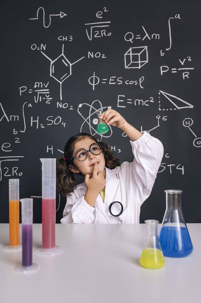 child scientist with glasses in lab coat looking a flask with chemical liquid, on blackboard background with science formulas, concept of back to school and successful female career, vertical photo - Fotoğraf, Görsel
