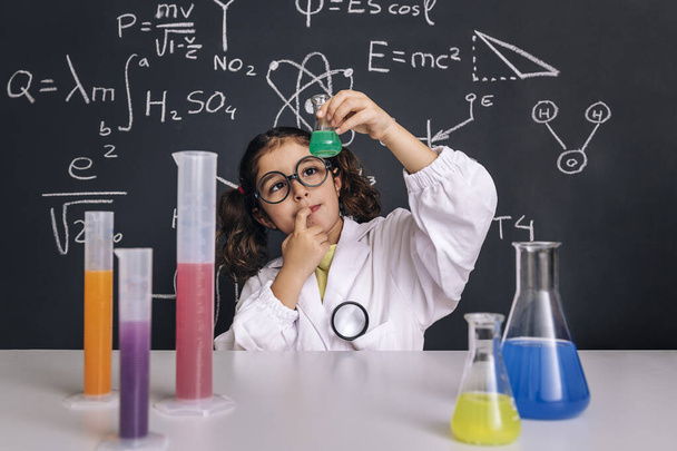 child scientist with glasses in lab coat looking a flask with chemical liquid, on blackboard background with science formulas, concept of back to school and successful female career - Photo, Image